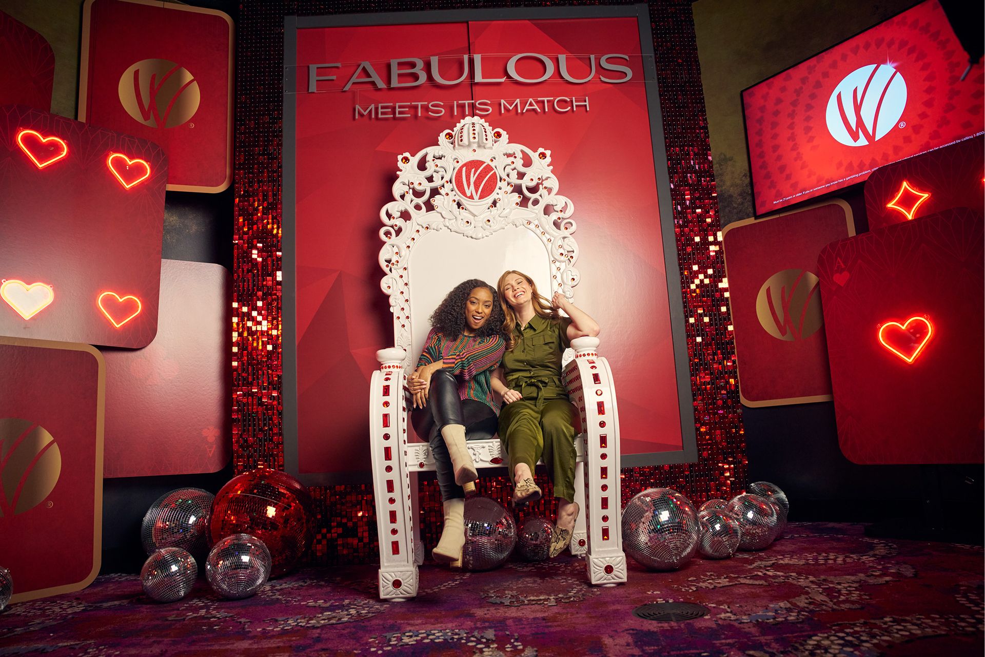 Two women sitting on a white ornate chair in front of a red, glittery backdrop with card suit designs and disco balls, with the text 'FABULOUS MEETS ITS MATCH' above them.